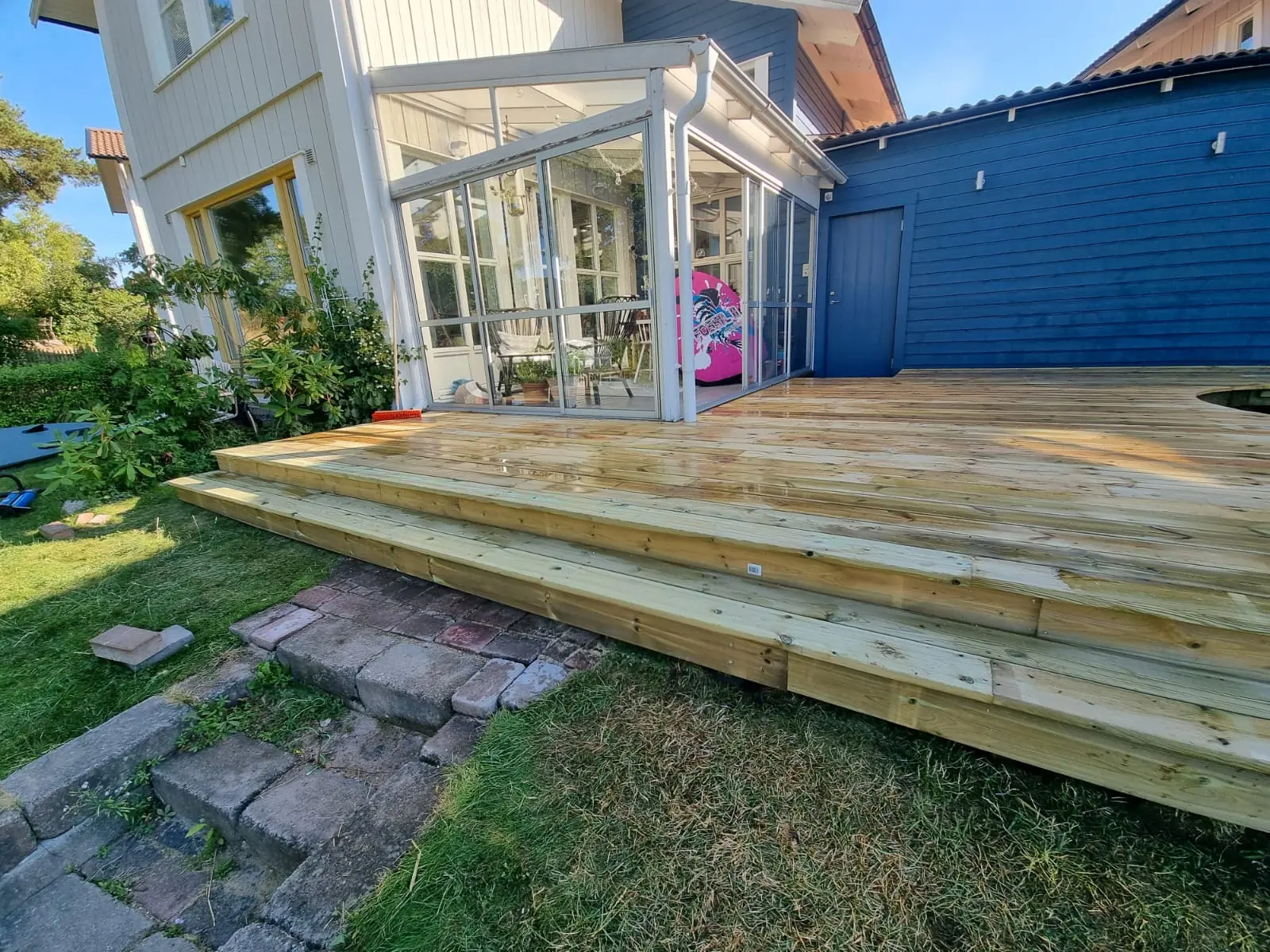 Uteplats i trä framför en inglasad veranda med blå fasad i Gustavsberg.